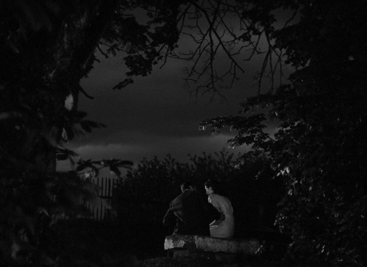 Krakatit (1948) movie still: wide — A dark night landscape featuring a pair of figures seated on a stone ledge. The scene is framed by dark tree…