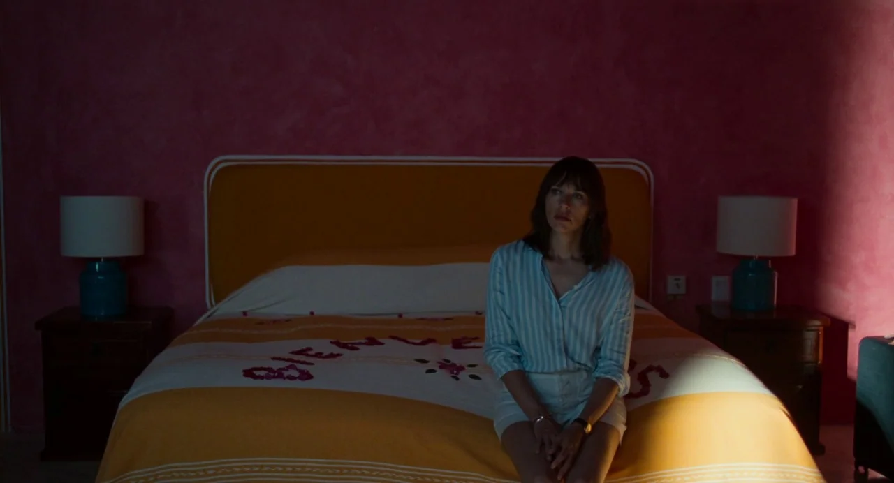 On The Rocks (2020) movie still: medium — A hotel room features vibrant pink walls and a yellow bedspread with flower petals. A woman sits alone on…