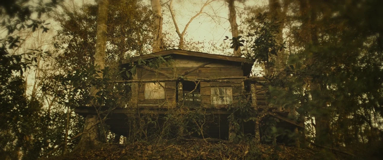 Blaze (2018) movie still: wide — A wooden treehouse with glass windows is nestled within a dense cluster of trees. Overgrown vegetation surrounds…