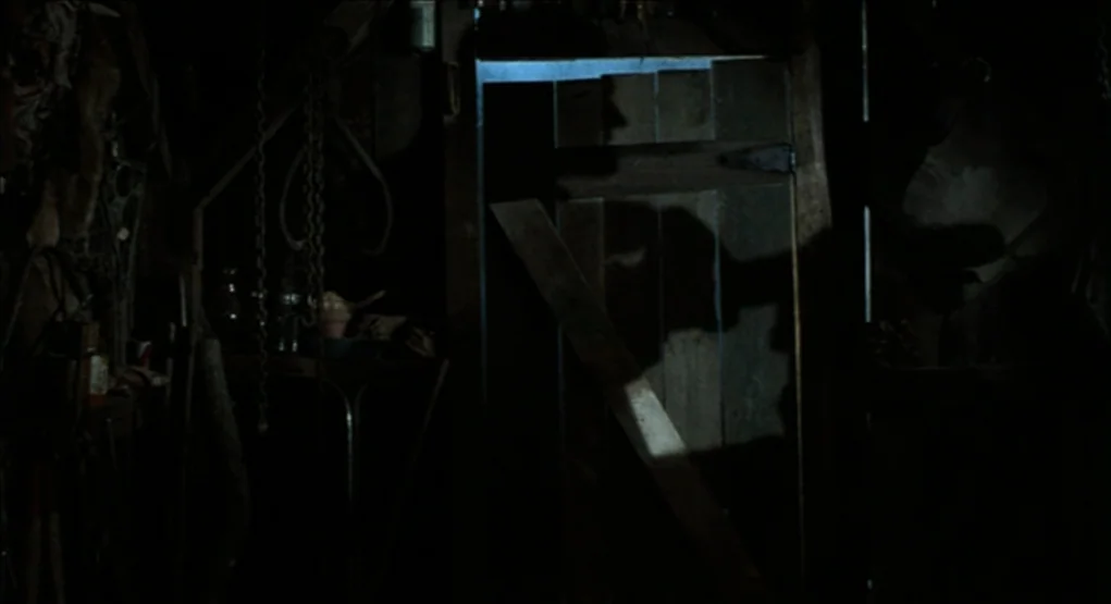 Evil Dead II (1987) movie still: medium — A dark workshop interior features cluttered tools and chains hanging on the left side. A distinct human…