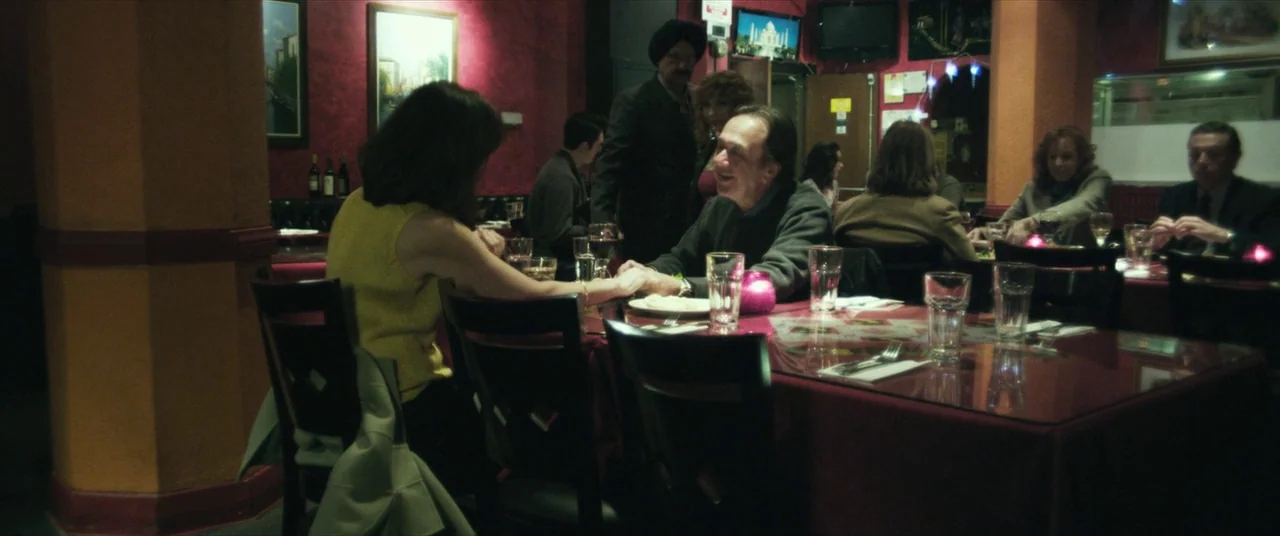 Maniac (2012) movie still: medium — A dimly lit restaurant features deep red textured walls and dark wooden furniture. A man and a woman hold hands…