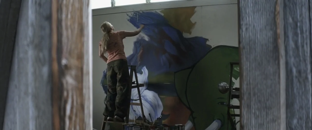 50 First Dates (2004) movie still: medium — A woman stands on a step ladder inside a studio space painting a mural. The mural depicts a large green…