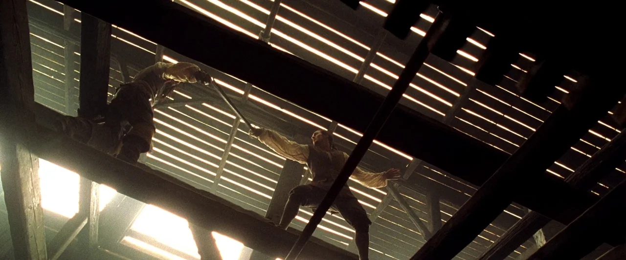 Pirates of the Caribbean: The Curse of the Black Pearl movie still: wide — Two men stand on wooden structural beams inside a building with a slat…