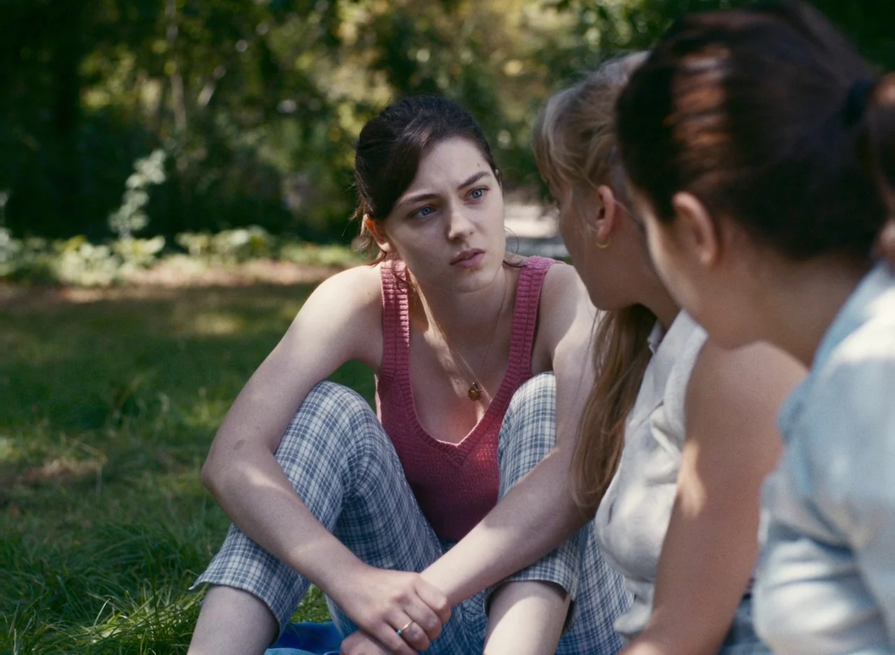 Happening (2021) movie still: medium — Three young women sit together on a patch of grass in a shaded park. The central woman faces the others while…