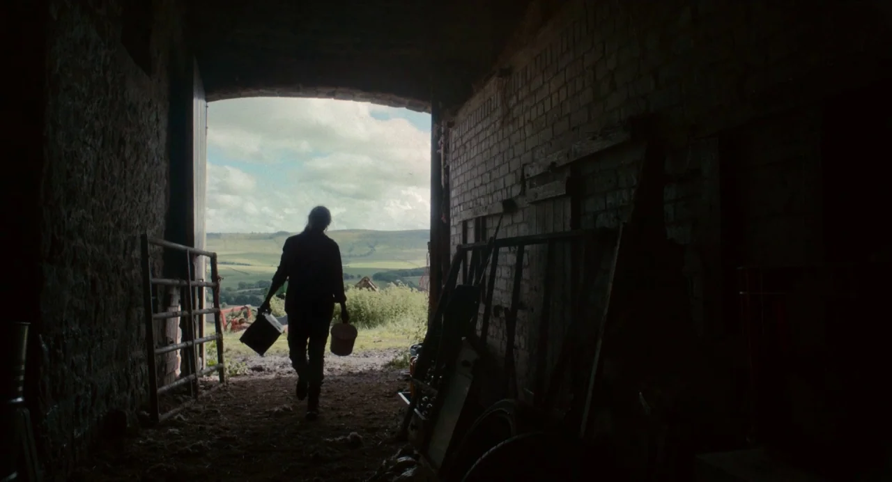 Dark River (2017) movie still: fullbody — A dark, brick-walled barn looks out onto a sunlit grassy hill under a cloudy sky. A person walks through…