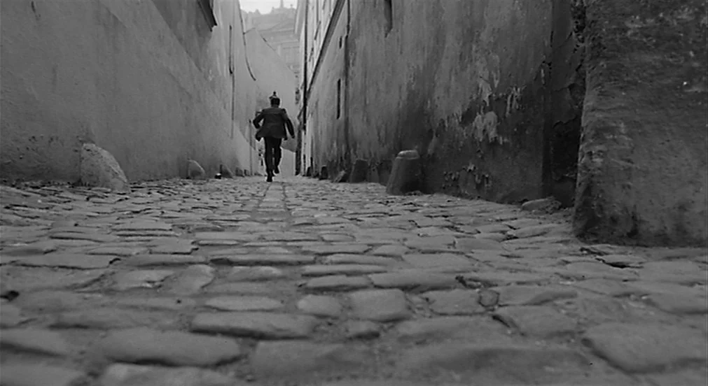 Kafka (1991) movie still: wide — A narrow stone-walled passage features a rough, uneven cobblestone ground surface. A man wearing a dark suit moves…