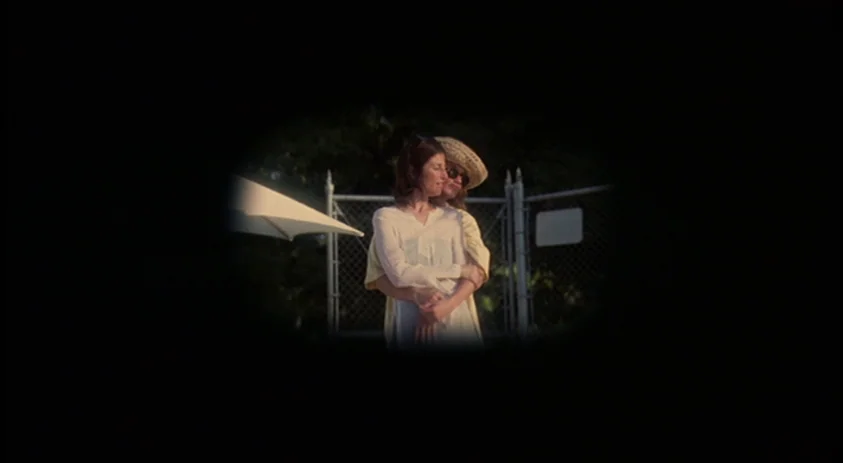 Being John Malkovich (1999) movie still: medium — Two women stand in front of a chain-link fence outdoors under an umbrella. One woman embraces the…