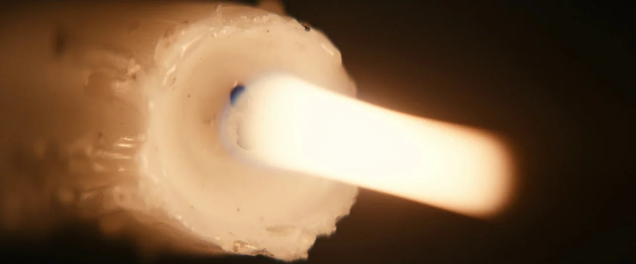 Leave The World Behind (2023) movie still: closeup — A close-up view of a burning candle wick centered within a wax cylinder. The flame extends…