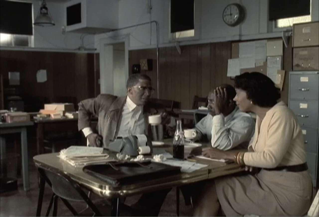 Boycott (2001) movie still: medium — A 1960s office setting features wood paneling, metal filing cabinets, and a laminate table. Three people are…