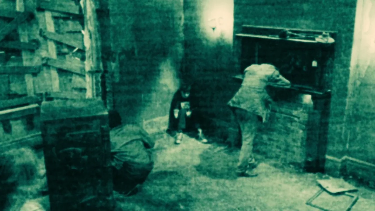 Saw II movie still: wide — An abandoned room with a boarded-up window features a brick fireplace along the wall. Three people are inside, one…