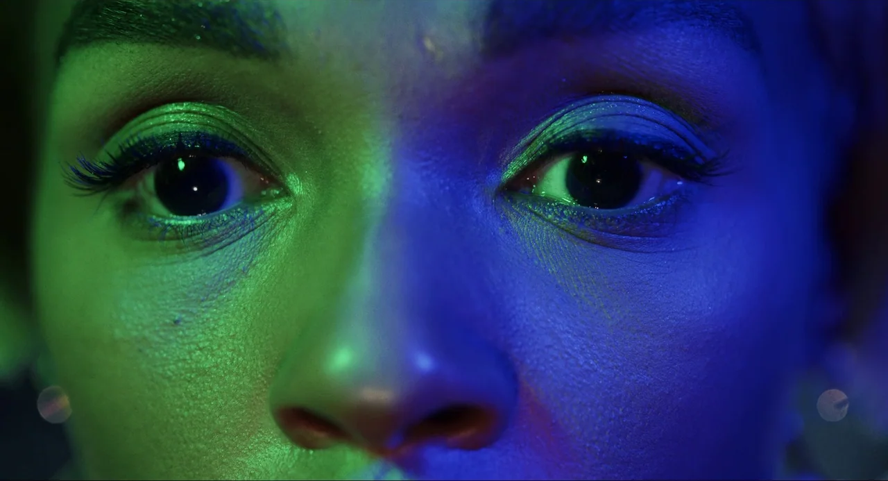 Glass Onion (2022) movie still: closeup — A close extreme portrait focuses on the eyes of a young woman. Intense neon green and blue light bisects…