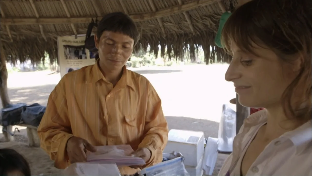 Aliens Inside Us (2013) movie still: medium — A man and a woman stand under a rustic thatched roof structure in an open area. The man in an orange…