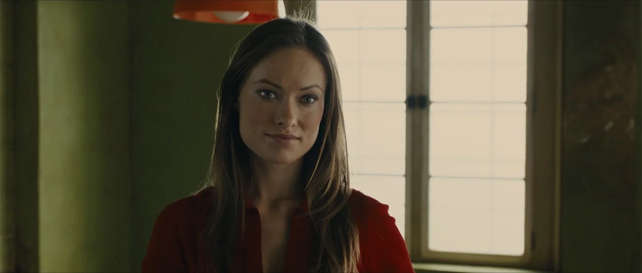In Time (2011) movie still: closeup — A room with muted green walls features a grid-patterned window that lets in daylight. A woman in a red shirt…