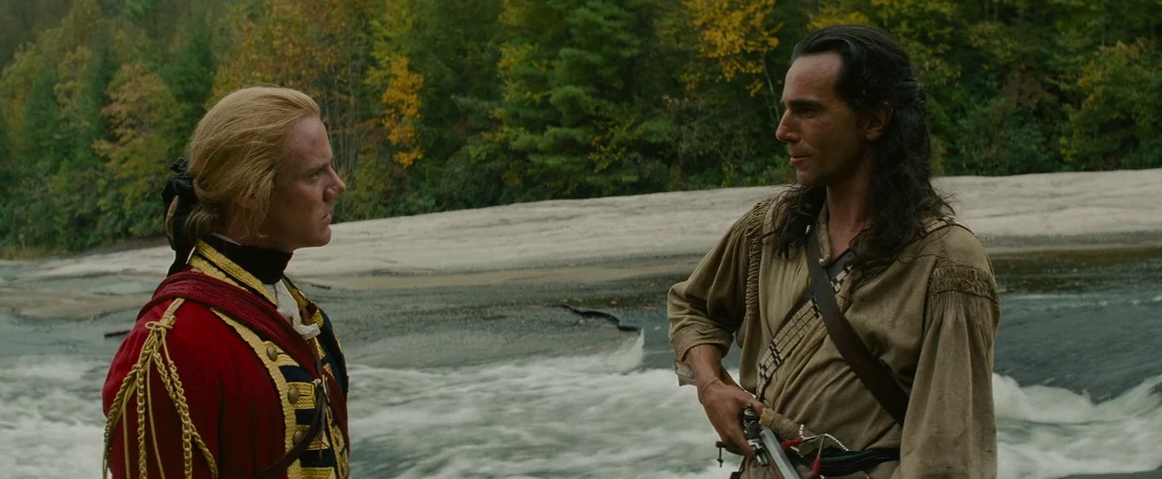 The Last of the Mohicans (1992) movie still: medium — A forested riverbank with a rushing stream and a light-colored rocky shoreline. A British…
