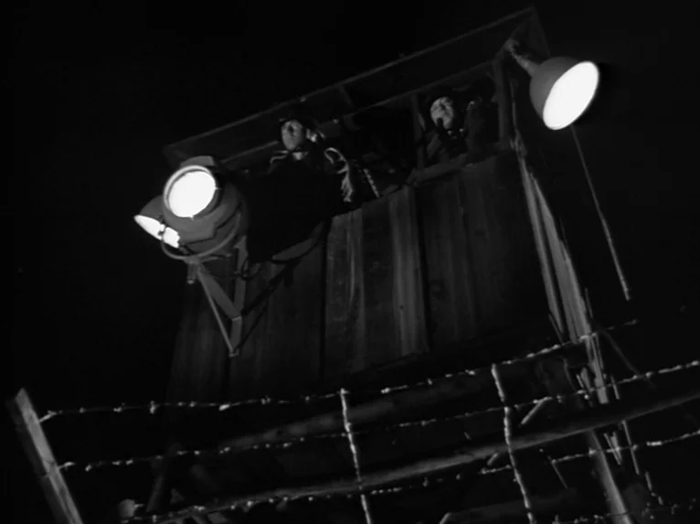 Stalag 17 (1953) movie still: wide — A tall wooden guard tower stands against a pitch black night sky. Two guards operate large spotlights while…