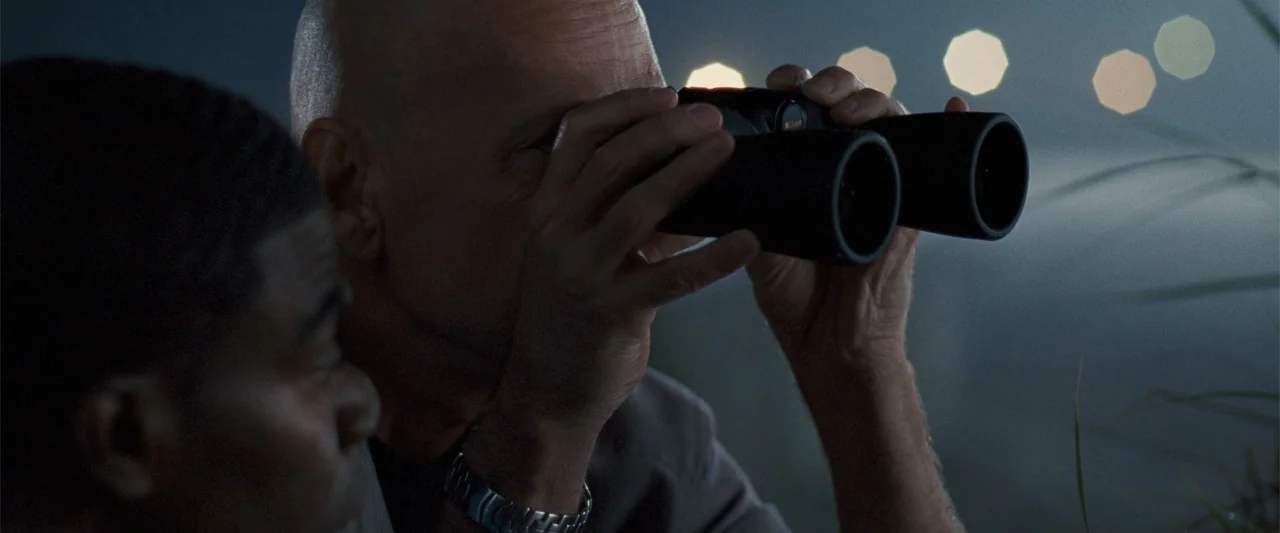 Cop Out (2010) movie still: closeup — A dark outdoor rooftop at night with out of focus lights in the distance. An older bald man peers through…