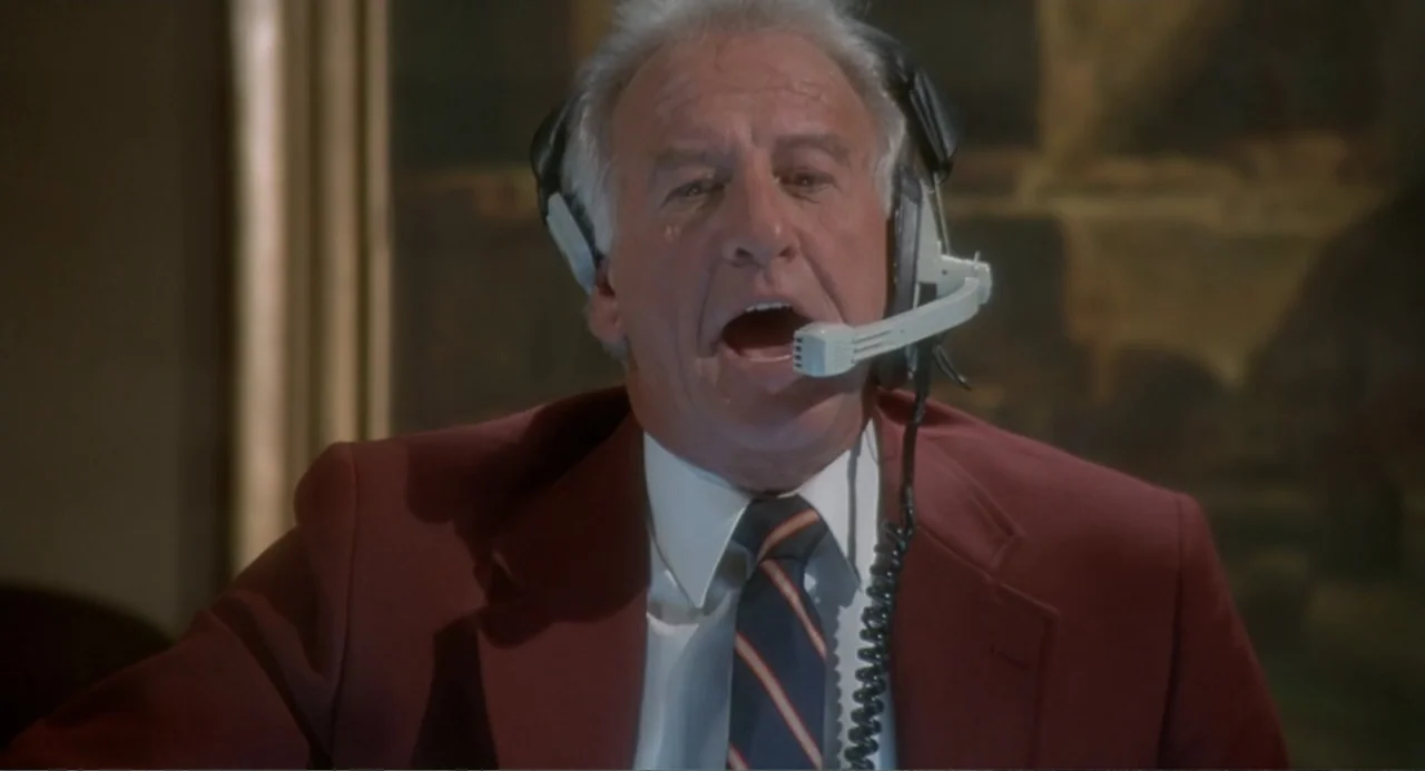 Fatal Instinct (1993) movie still: closeup — An interior broadcast booth features a dark wall backdrop and a framed picture. An elderly man in a…