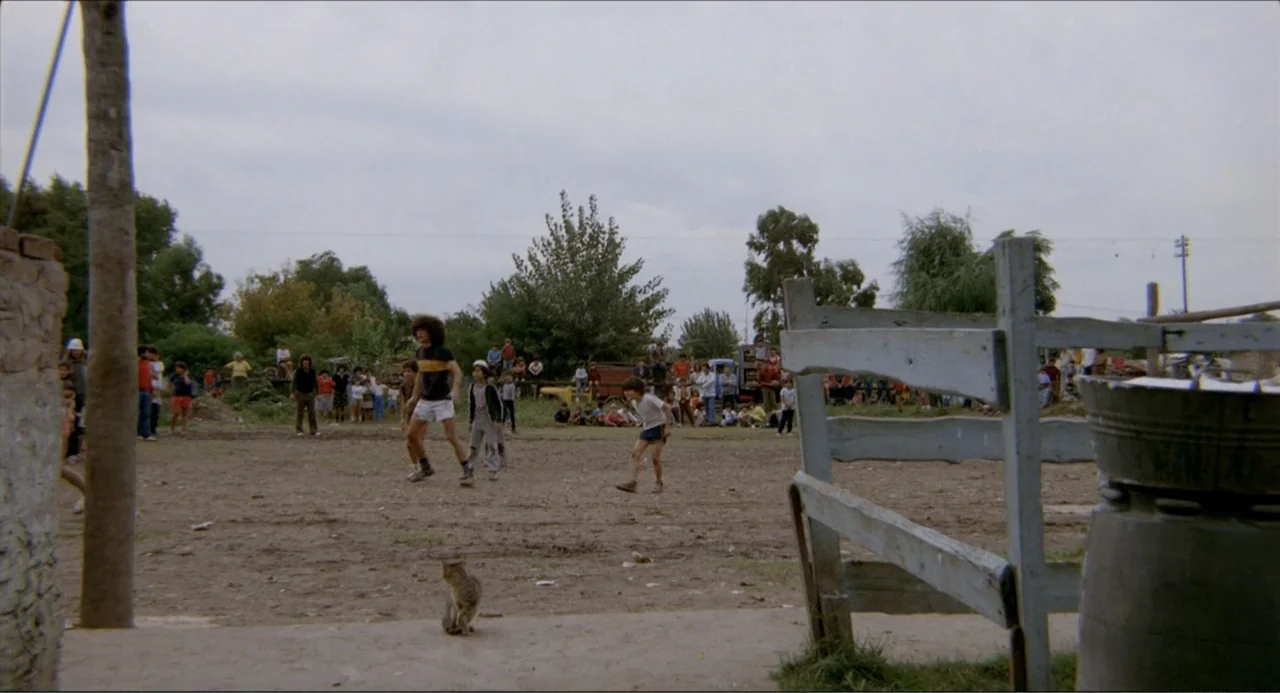 Waiting for the Hearse (1985) movie still: wide — A rural dirt sports field is surrounded by trees and a sparse crowd. Several children play soccer…