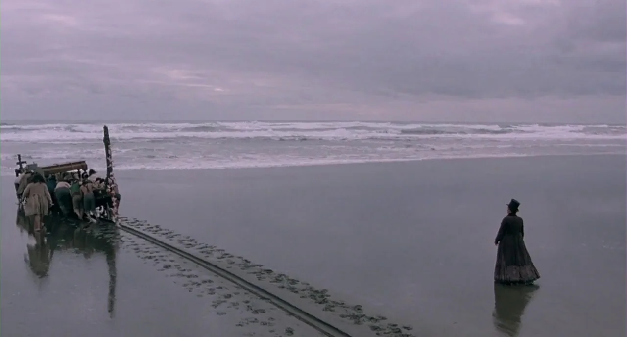 The Piano (1993) movie still: wide — A wet, overcast beach stretches towards the churning surf under a grey, clouded sky. A lone woman in…