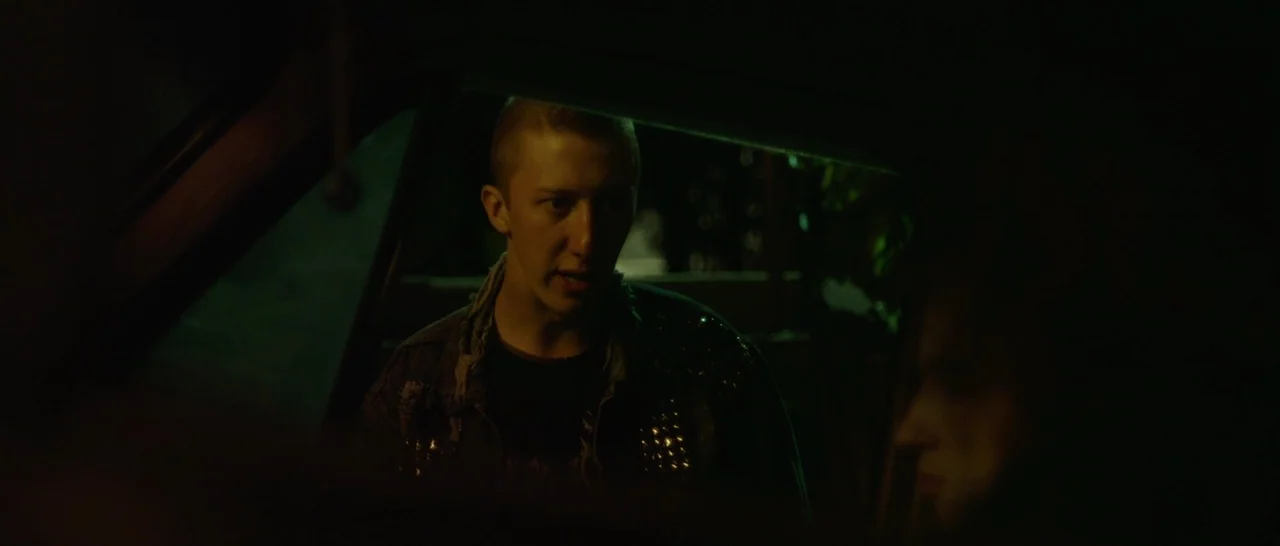 Green Room (2015) movie still: medium — A man sits inside a dark vehicle at night while illuminated by green light. The man talks to an unseen…