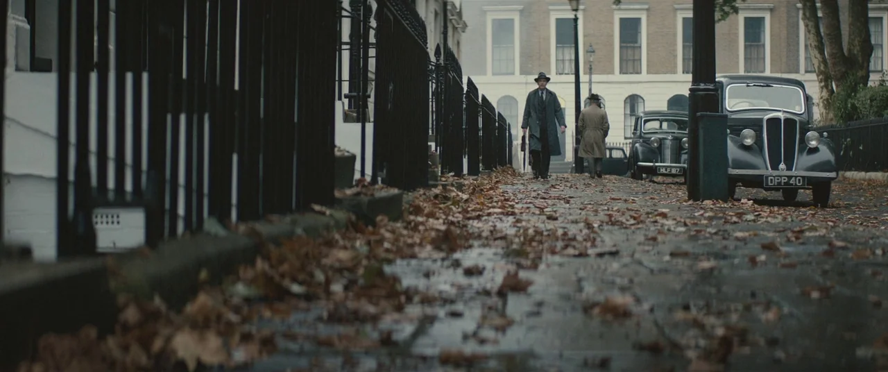Christopher Robin (2018) movie still: wide — A residential London street features neoclassical townhouses and vintage cars parked along the curb.…