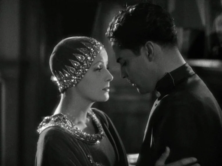 Mata Hari (1931) movie still: medium — An ornate interior room with dark, out-of-focus architecture serves as the backdrop. A woman in a sequined…