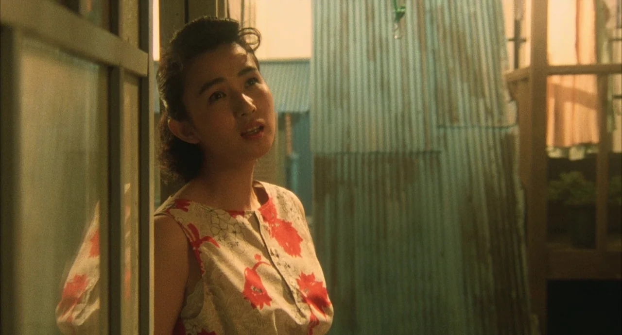 The Discarnates (1988) movie still: medium — A narrow outdoor space features corrugated metal walls and a window frame. A young woman stands looking…