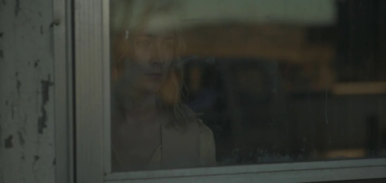 Foe (2023) movie still: closeup — A woman stands behind a window pane at night. She looks forward while her face is reflected on the glass surface.