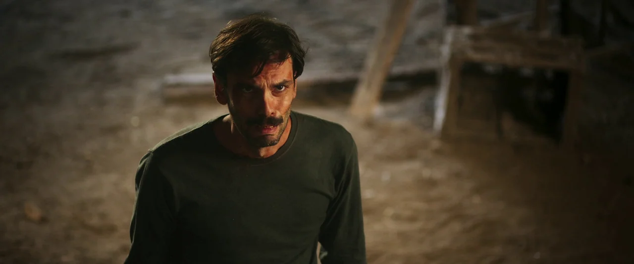 When Evil Lurks (2023) movie still: medium — An open dirt lot with a blurred wooden structure in the background. A man stands in the dirt looking…
