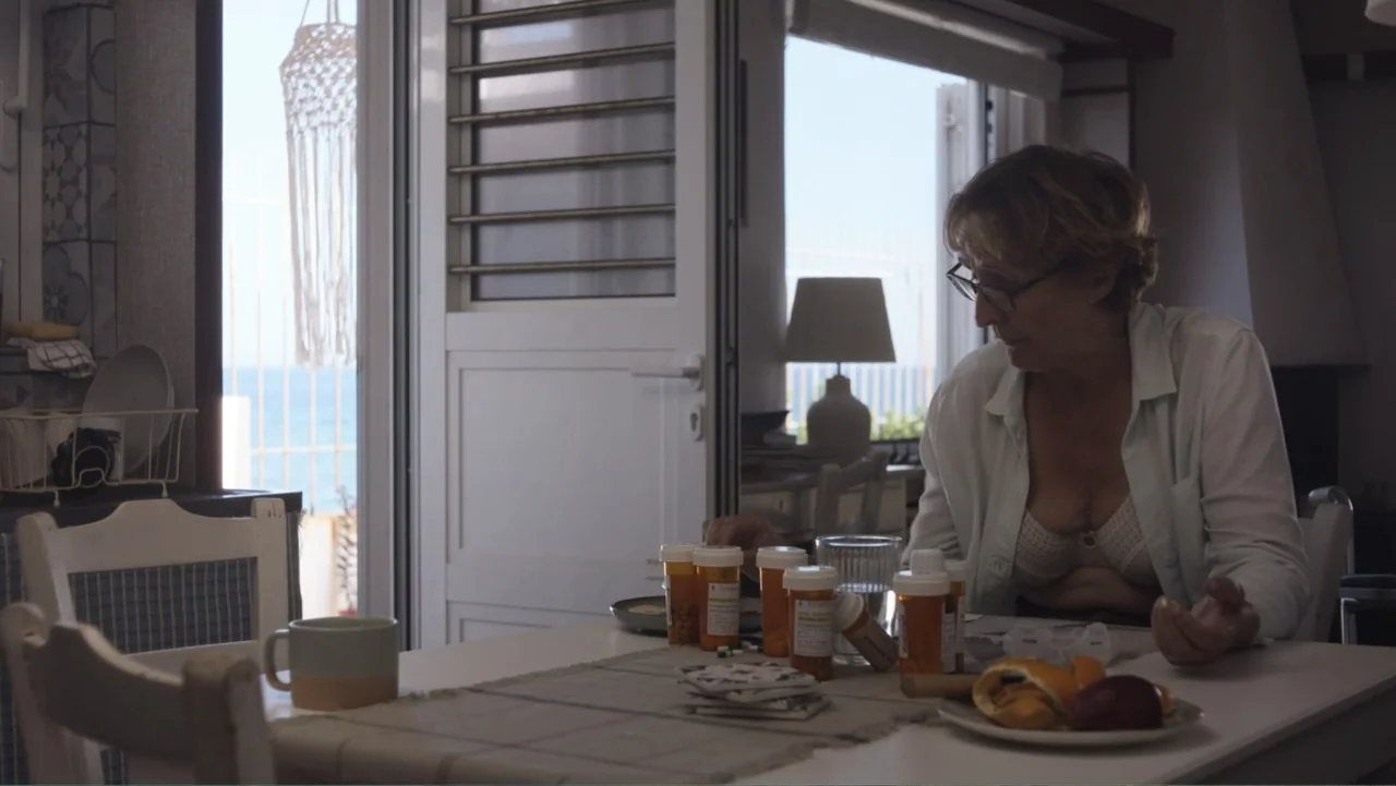 Hot Milk (2025) movie still: medium — A sunlit dining room features a table covered with various prescription medication bottles. An older woman in…