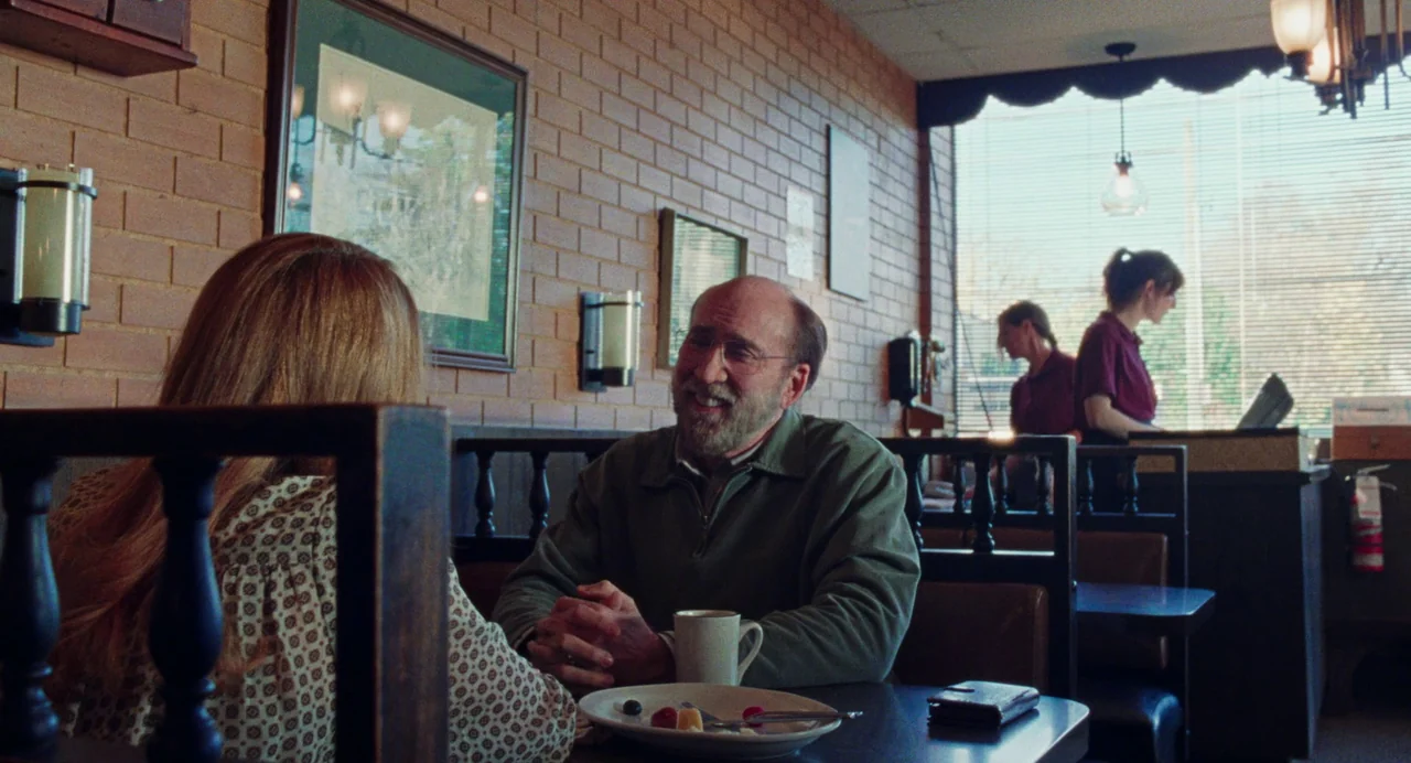 Dream Scenario (2023) movie still: medium — A diner booth with a tan brick wall and wooden dividers separates the tables. A man sits across from a…