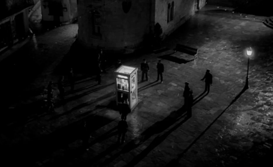 Vivement Dimanche (1983) movie still: wide — An illuminated telephone booth sits in the center of a dark stone-paved plaza at night. Several men in…
