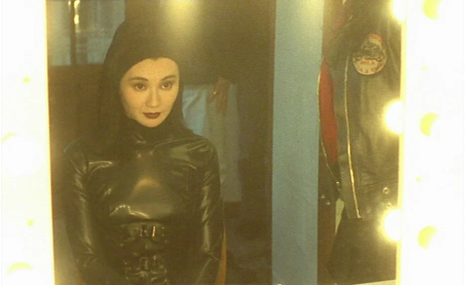 Irma Vep (1996) movie still: medium — A woman stares into a brightly lit dressing room vanity mirror. Her reflection is visible against a plain…