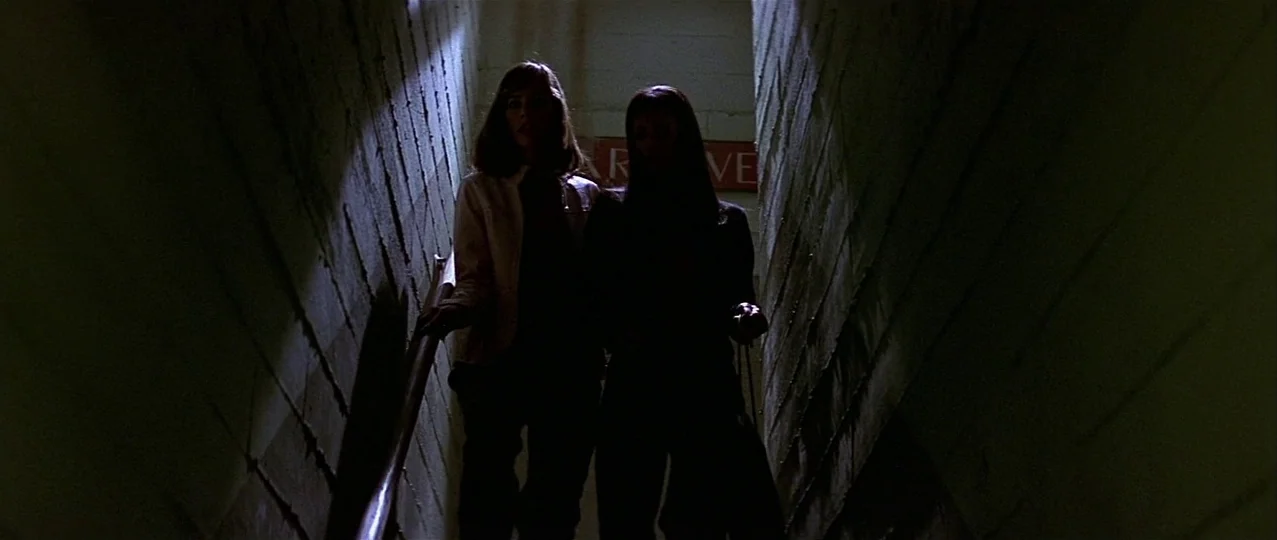 Scream 3 (2000) movie still: medium — Two women walk down a narrow concrete stairwell with rough block walls. A red sign is partially visible on the…