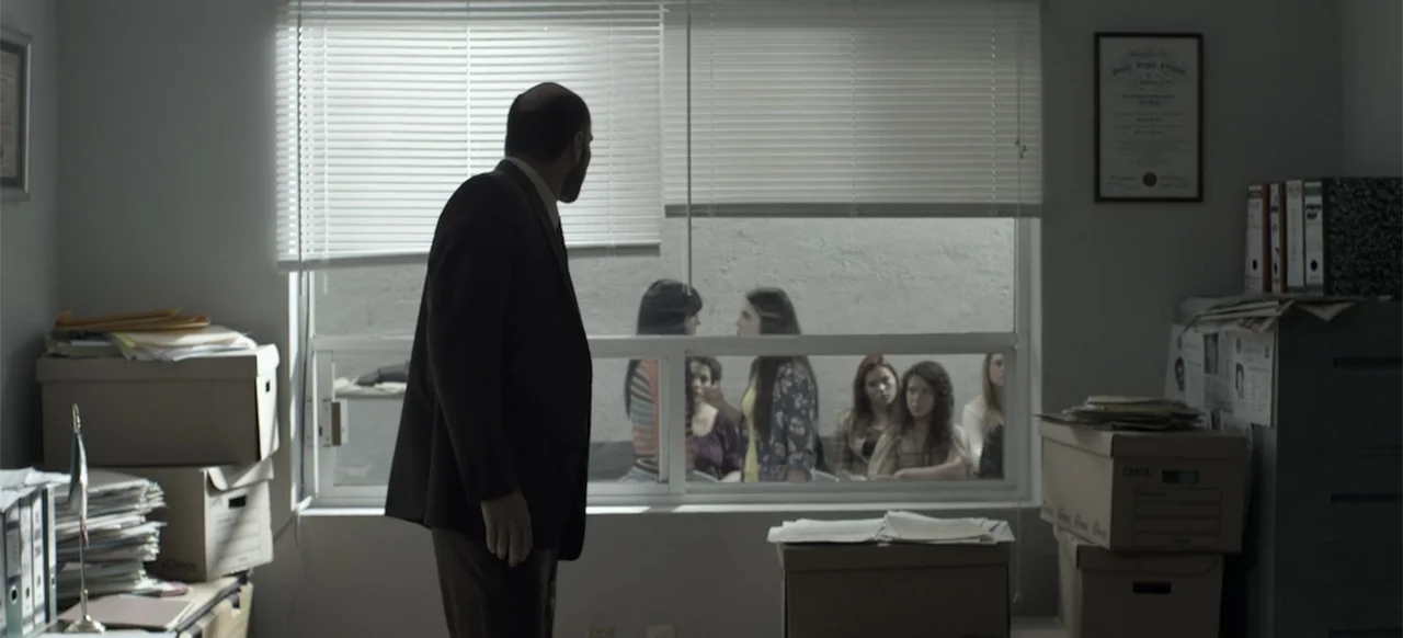 About the Deaths (2016) movie still: medium — An interior office space features stacked cardboard boxes and paperwork on desks. A man looks through…