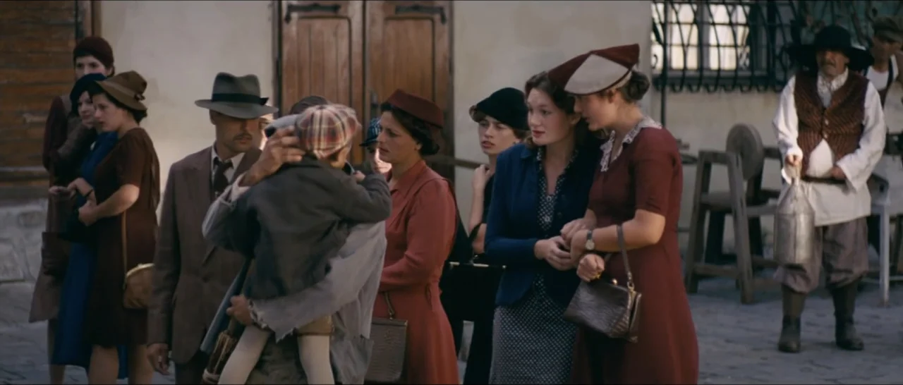 A Farewell to Fools (2013) movie still: medium — A group of people dressed in 1940s attire stand in a dusty outdoor square. A man holds a child…