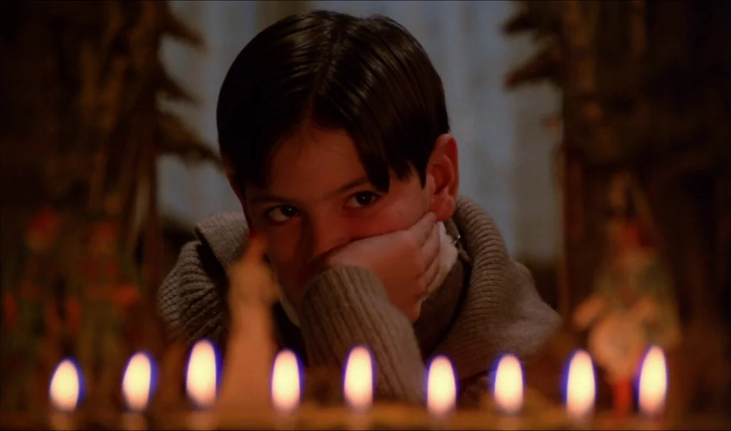 Fanny and Alexander (1982) movie still: closeup — A dark indoor space is dimly lit by a row of small flickering candles. A young boy rests his chin…