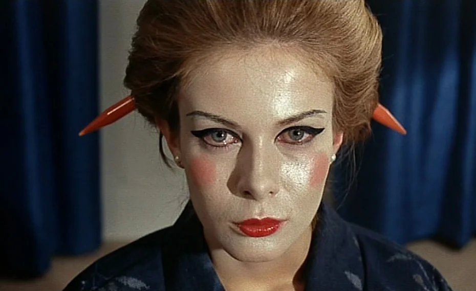 Bed & Board (1970) movie still: closeup — A woman stares forward against a background of solid dark blue curtains. She wears heavy white face makeup…
