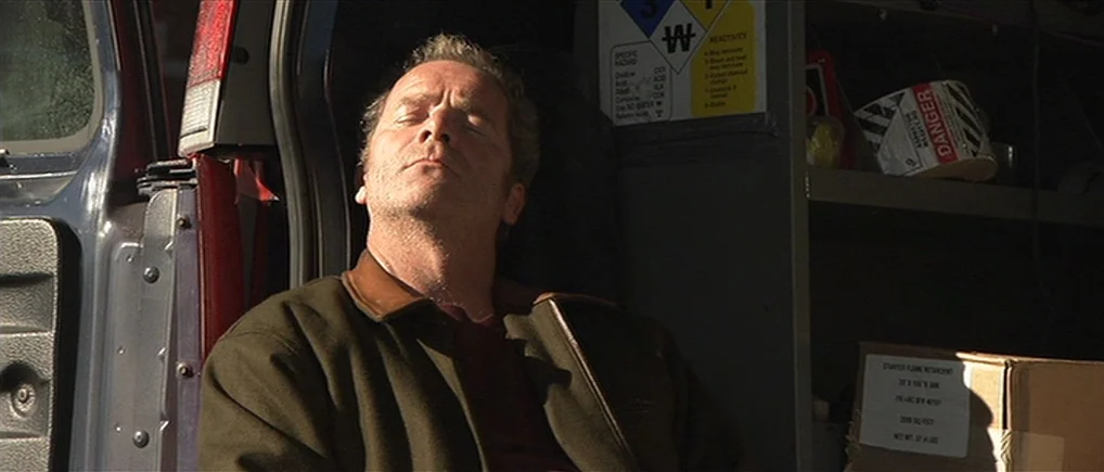 Session 9 (2001) movie still: medium — A dark cargo area of a van contains hazard warning signs and cardboard boxes. A man sits inside with eyes…