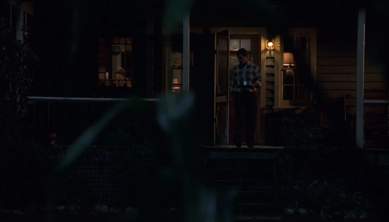Signs (2002) movie still: wide — The front porch of a wood-sided house at night features a glowing entry light. A man steps out of the open doorway…