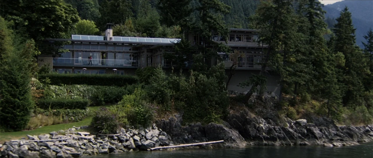 Elektra (2005) movie still: establishing — A modern multi-story glass-fronted house sits perched on a rocky shoreline amidst dense pine trees. The…