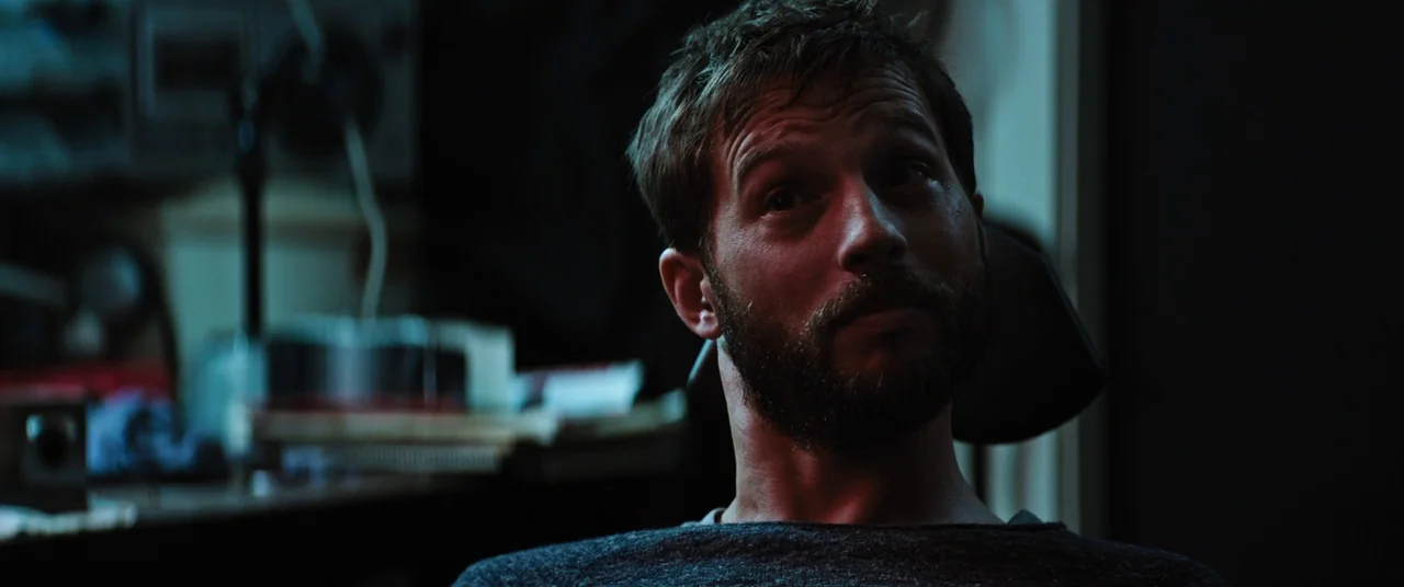 Upgrade (2018) movie still: closeup — A dark workspace with equipment visible on a blurred desk in the background. A man with a beard stares towards…