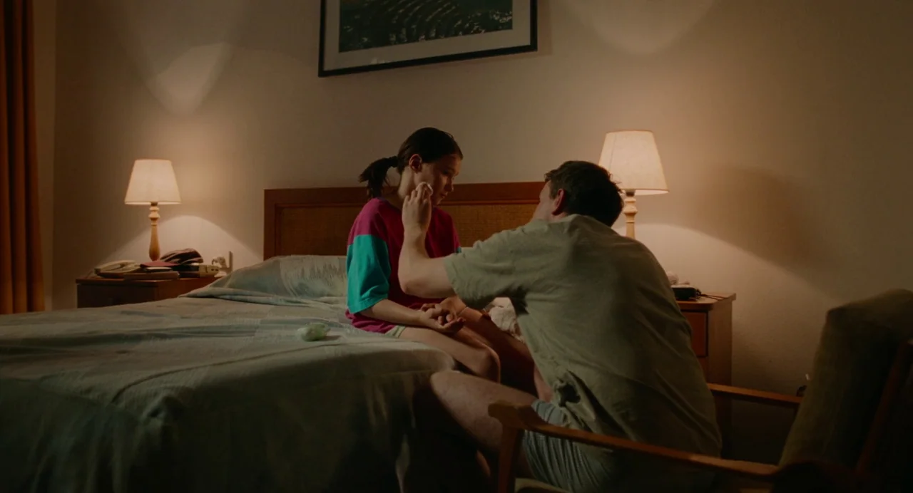 Aftersun (2022) movie still: medium — A standard hotel room features a bed with blue patterned bedding and two lamps. An adult man sits on a chair…