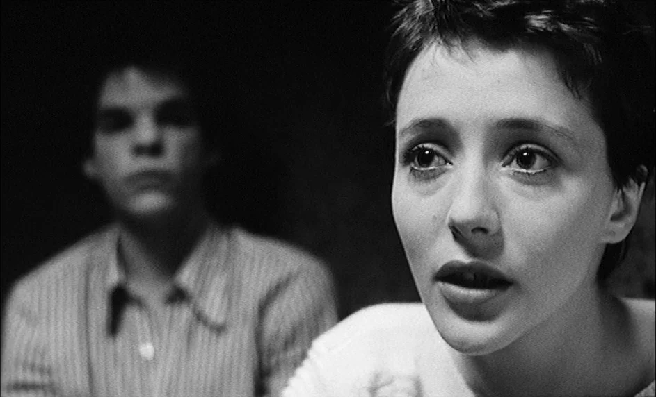 Boy Meets Girl (1984) movie still: closeup — A dark indoor space where the foreground subject is illuminated against a black background. A woman in…