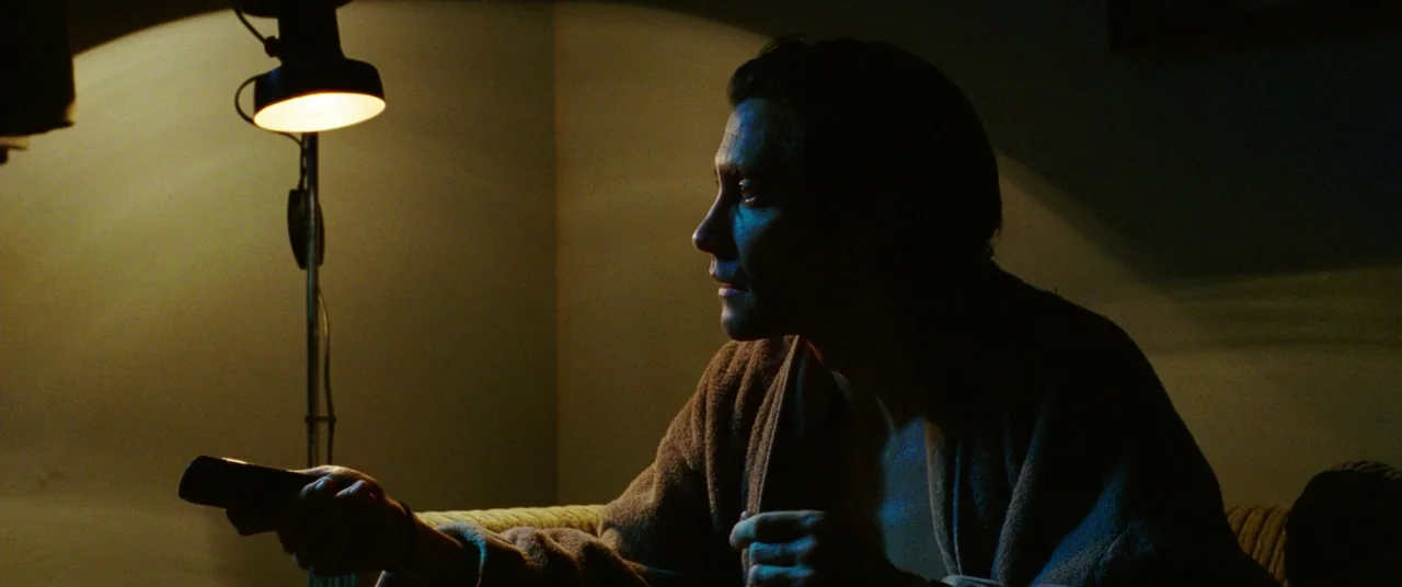 Nightcrawler (2014) movie still: medium — A dark, dimly lit indoor space with a plain wall and minimal decoration. A man in a bathrobe holds a…