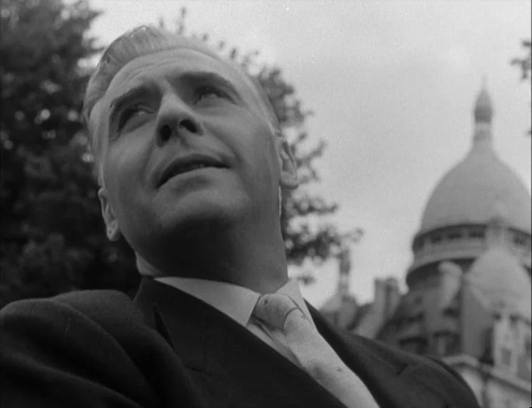 Bob le Flambeur (1956) movie still: closeup — A man in a suit stands in an urban outdoor space with trees. The dome of a large church is visible in…