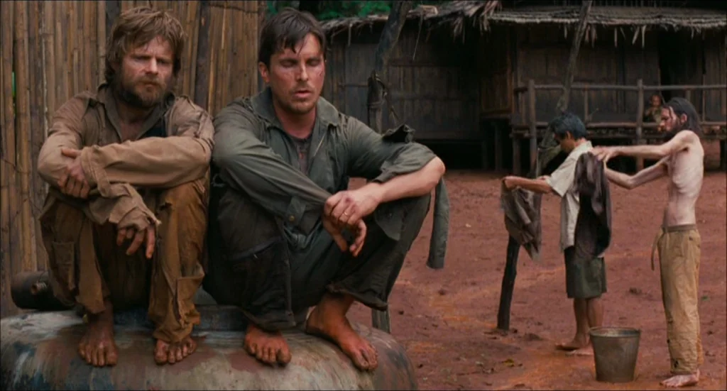 Rescue Dawn movie still: fullbody — A rural encampment with bamboo structures and red dirt ground under overcast daylight. Two men sit on a water…