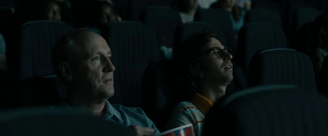 Brigsby Bear (2017) movie still: medium — A dark movie theater interior filled with rows of empty and occupied plush seats. Two men in the middle…