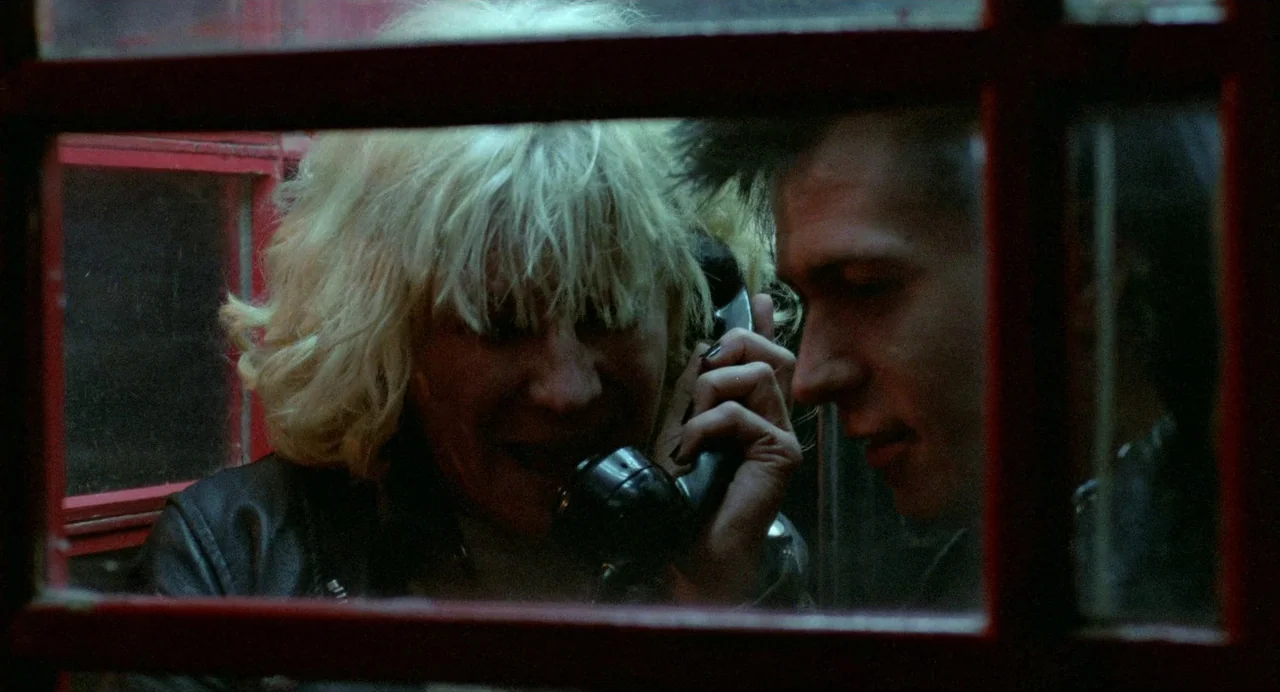 Sid and Nancy (1986) movie still: medium — Two individuals occupy a confined red phone box at night. The blonde woman holds a receiver to her ear…