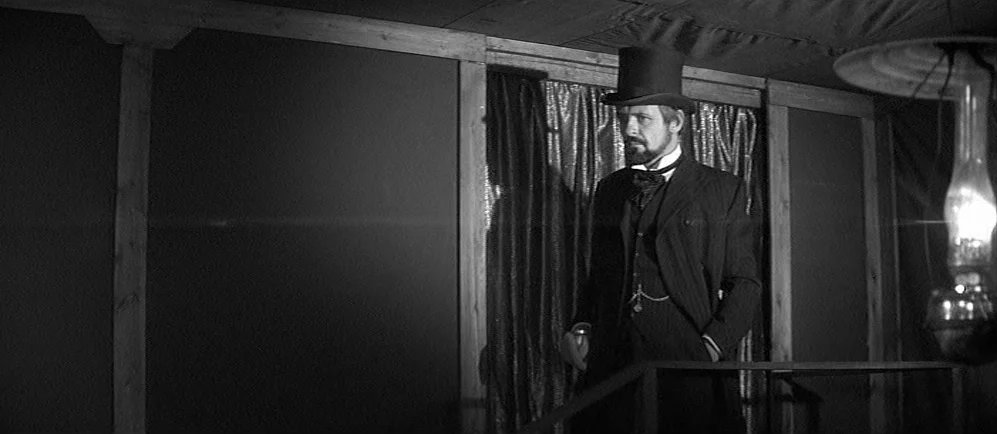 The Elephant Man (1980) movie still: medium — An interior space features wood support beams and dark curtains against the back wall. A man dressed…